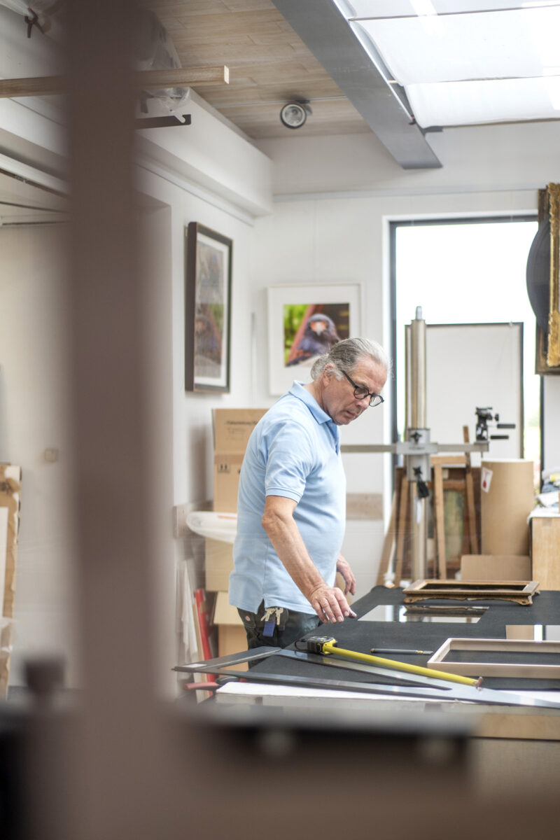 Arbeitsfoto von Rahmenbauer Joachim Konradi in seiner Werkstatt im Bergischen Land. Zu sehen ist Joachim Konradi beim Zuschneiden von Glas. Er hat graues, etwas längeres Haar zu einem Zopf gebunden und trägt ein hellblaues Polohemd. Er schaut nach unten auf seine Arbeit.