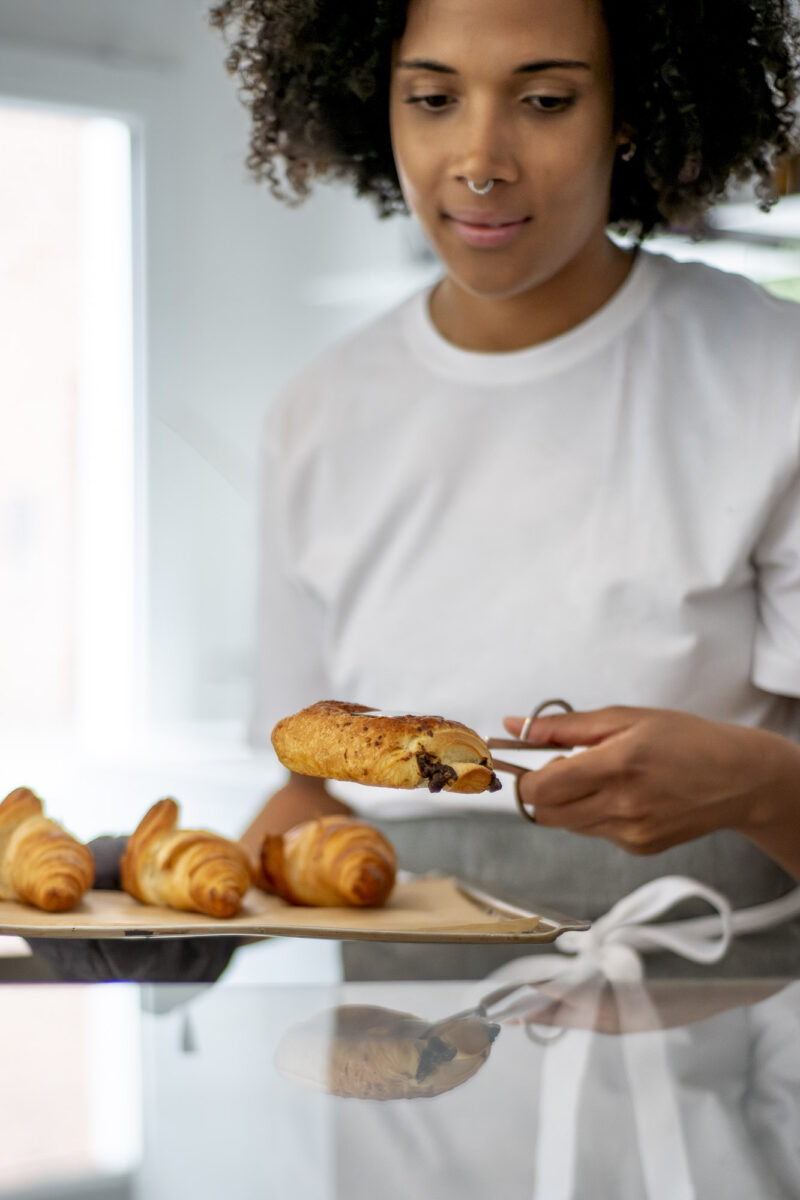 Foto von einer jungen Frau mit dunklen, lockigen Haaren, die ein Tablett mit Croissants, die sie gerade in eine Auslage einsortieren möchte, in der Hand hält. Sie trägt ein weißes T-Shirt und eine Schürze.