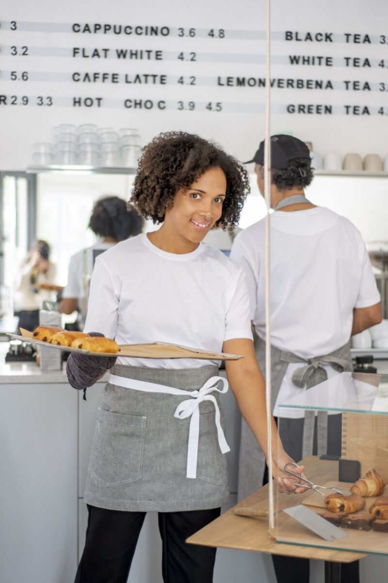 Foto von einer Frau mit dunklen, lockigen Haaren in einem modernen Café, die ein weißes T-Shirt und eine Schürze trägt und ein Tablett mit Schoko-Croissants in der Hand hält. Dabei schaut und lächelt sie in die Kamera.