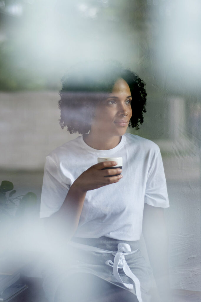 Foto von einer jungen Frau mit dunklen, lockigen Haaren beim Kaffee trinken durch eine Fensterscheibe fotografiert mit Spiegelungen. Die Frau schaut versonnen und lächelnd zur Seite. Sie trägt ein weißes T-Shirt.