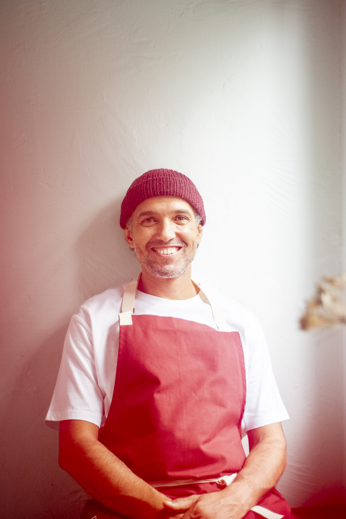 Portrait von einem Mann mittleren Alters mit leicht gräulichen Haaren und Mütze, der vor einer weißen Wand sitzt und lächelnd in die Kamera schaut. Er trägt ein weißes T-Shirt und eine rote Servier-Schürze. Von der Seite beleuchtet diffuses Sonnenlicht sein Gesicht. Er sieht zufrieden und happy aus.