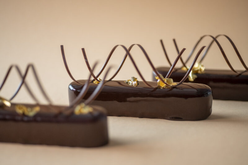 Produktaufnahmen von braunen, mit Blattgold kunstvoll verzierten, länglichen Schokoladen-Törtchen vor einem beigen Hintergrund.