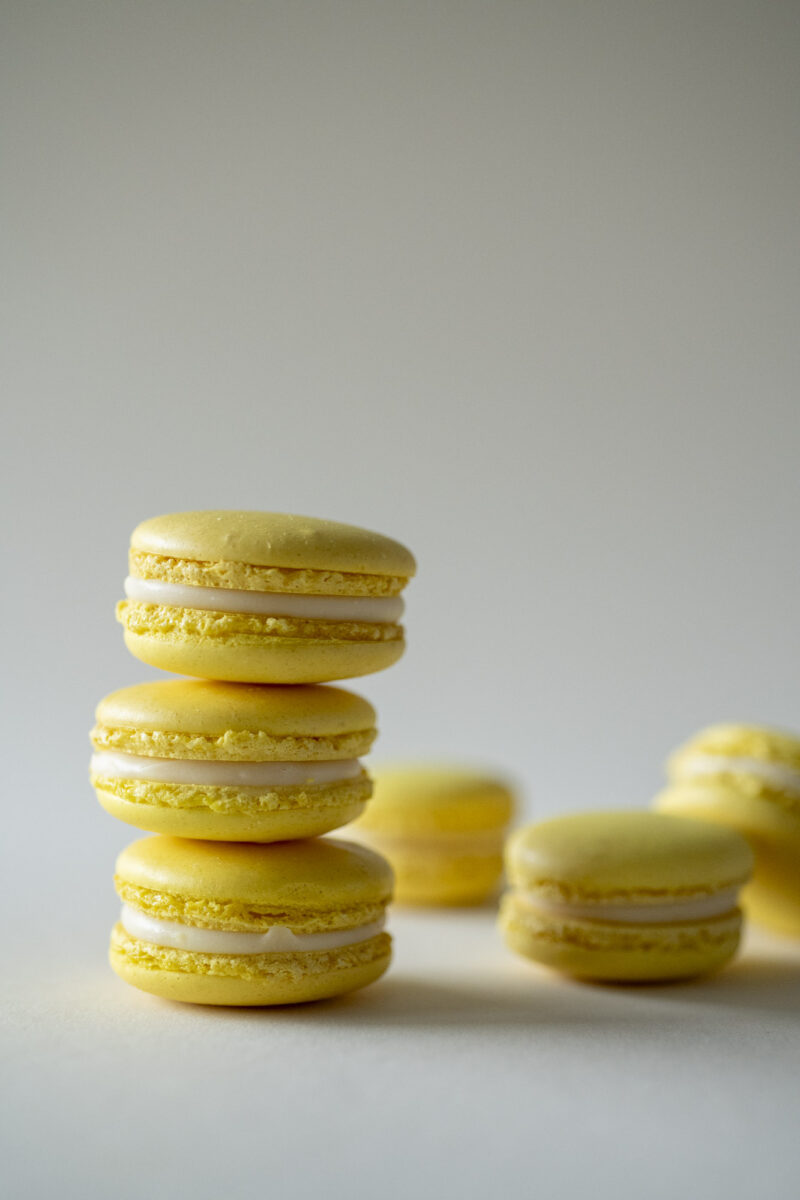 Produktaufnahme von aufeinander gestapelten gelben Maccarons vor einem gräulichen Hintergrund. Die Maccarons sehen sehr schmackhaft aus.