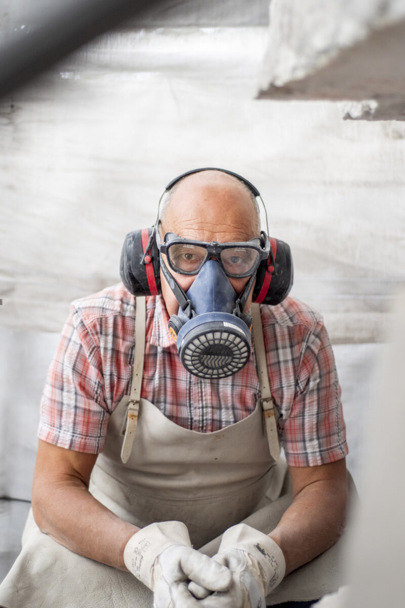 Foto von einem älteren Mann, der auf einer Baustelle sitzt mit Atemmaske und Gehörschutz. Er trägt Arbeitskleidung, Arbeitshandschuhe und ein kariertes Hemd. Er schaut in die Kamera und sein Gesichtsausdruck ist ernst.