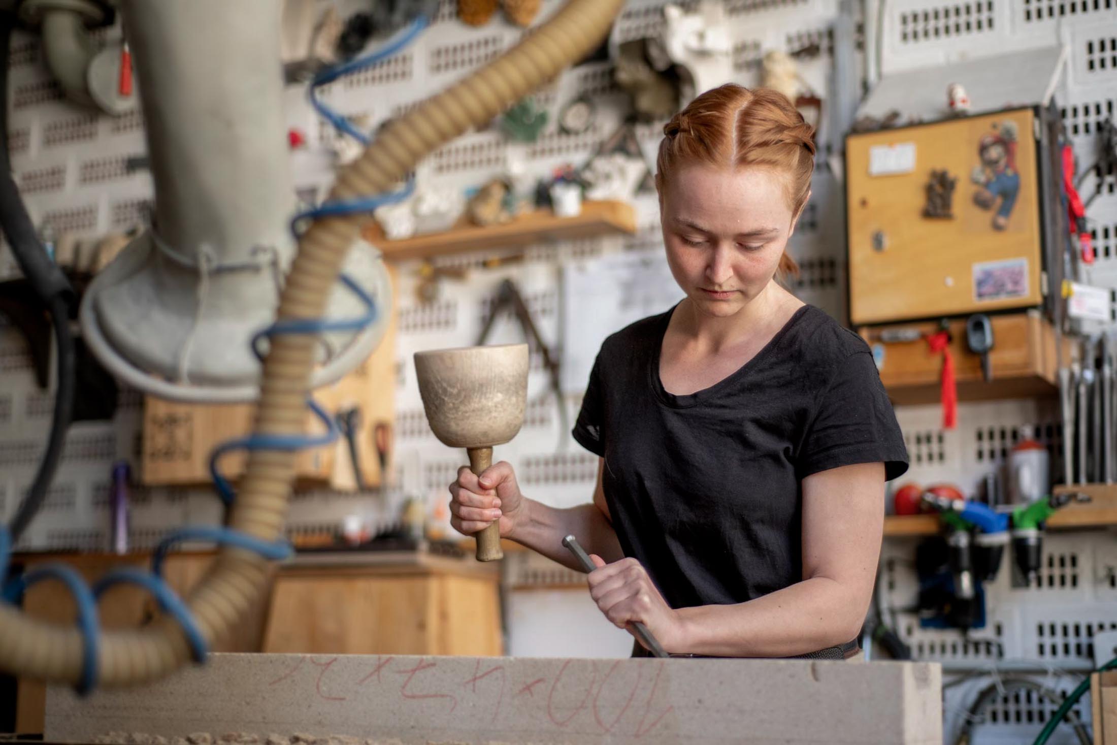 Foto von einer jungen Steinmetzen mit rötlichem Haar, die mit konzentriertem Gesichtsausdruck in der Werkstatt einen Stein mit Hammer und Meißel bearbeitet.