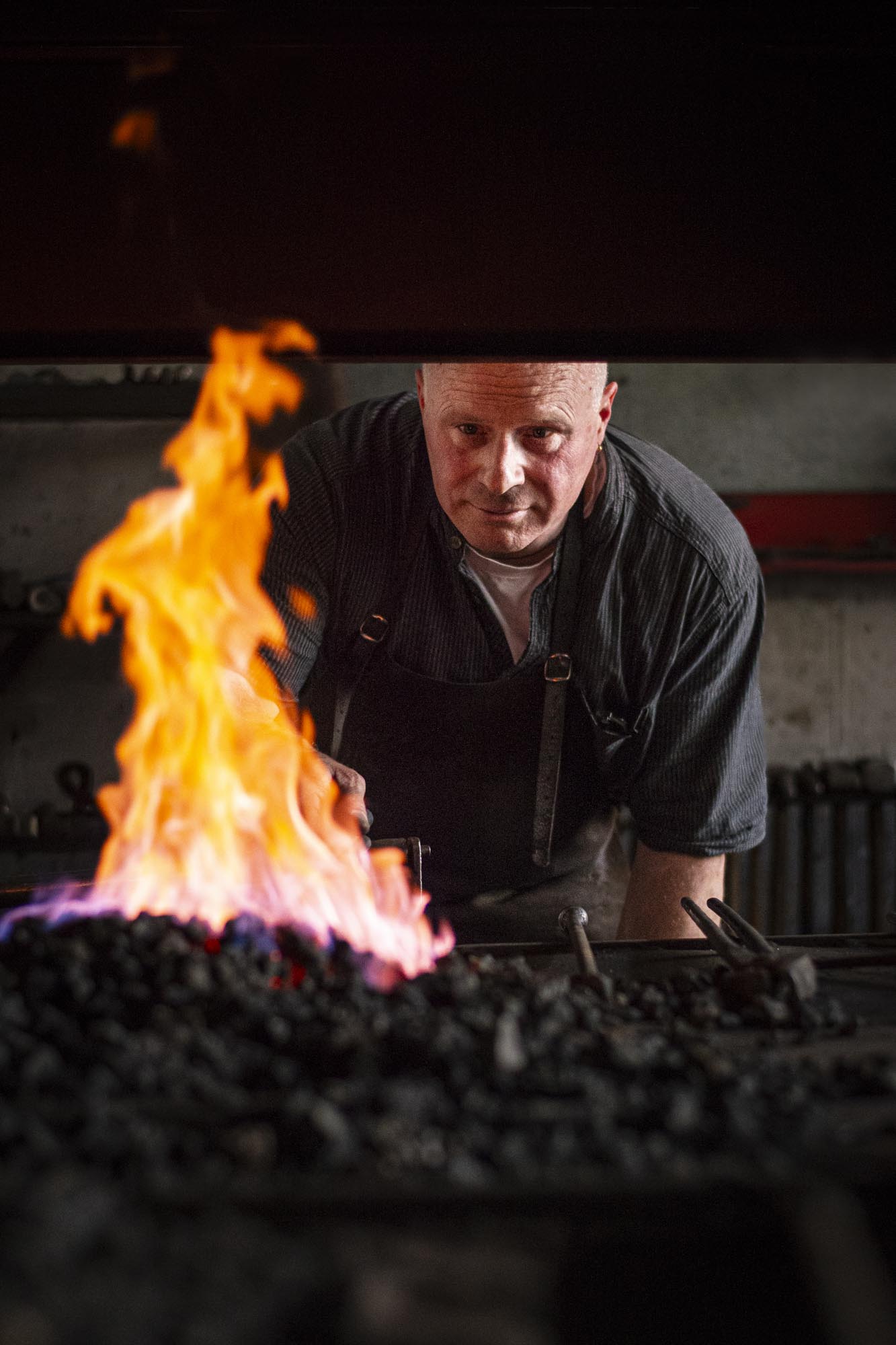 Schmied bei der Arbeit in der Werkstatt der Dombauhütte des Kölner Doms. Ein Mann in dunkler Schürze beugt sich konzentriert über die offene Glut, während die Flammen hell lodern. Das Bild zeigt die Kraft des Feuers, die Präzision des Handwerks und die traditionsreiche Arbeit, die hinter der Erhaltung historischer Baukunst steckt.
