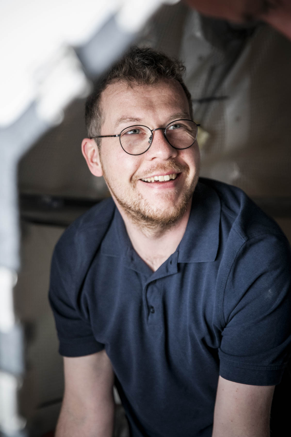 Porträtaufnahme: Ein junger Glaser mit Brille und dunkelblauem Polohemd auf einem Gerüst an einem der Domfenster im Innenraum des Kölner Doms. Er schaut zur Seite und lächelt.