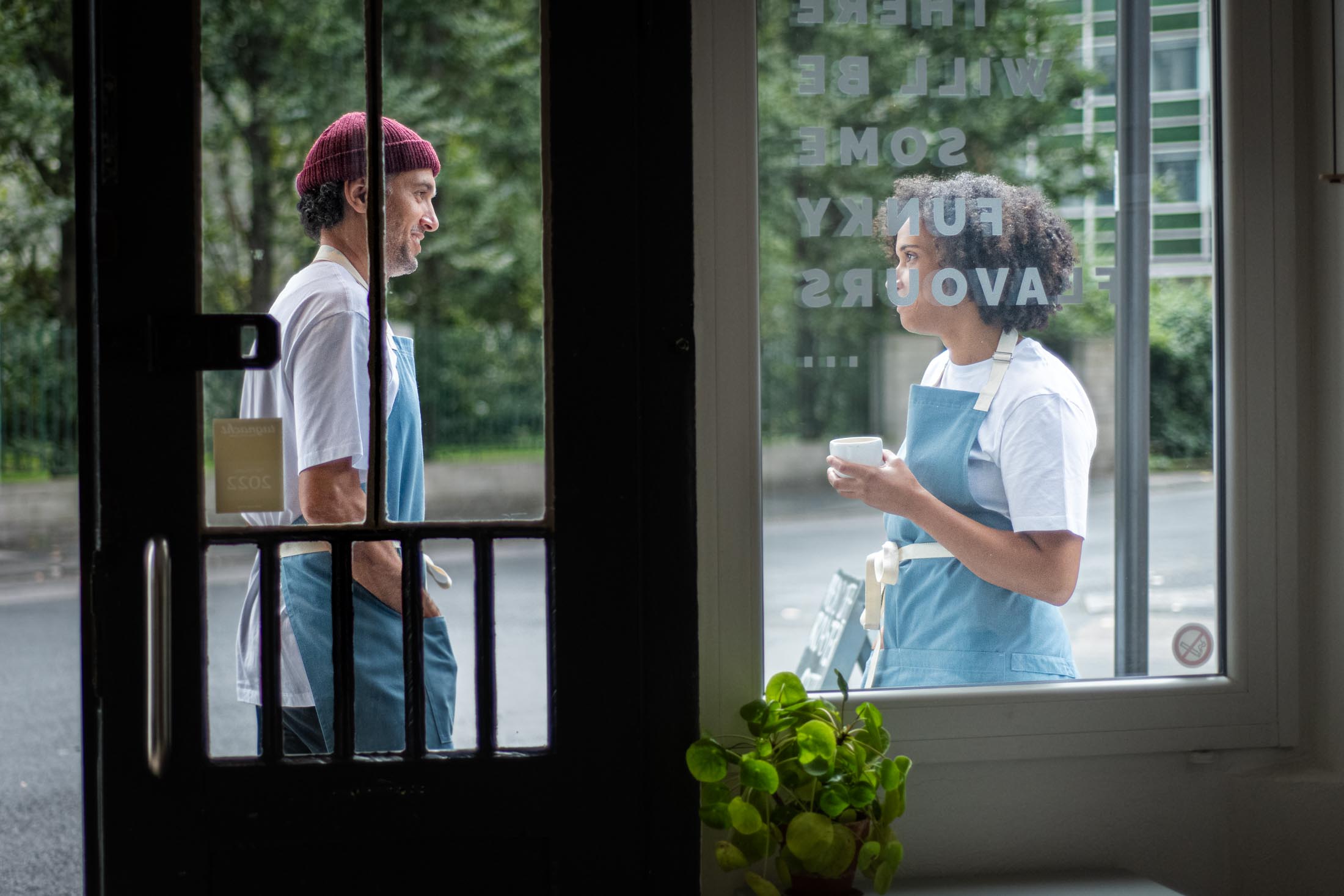 Zwei Personen in Gastro-Arbeitsbekleidung durch ein Caféfenster von innen heraus